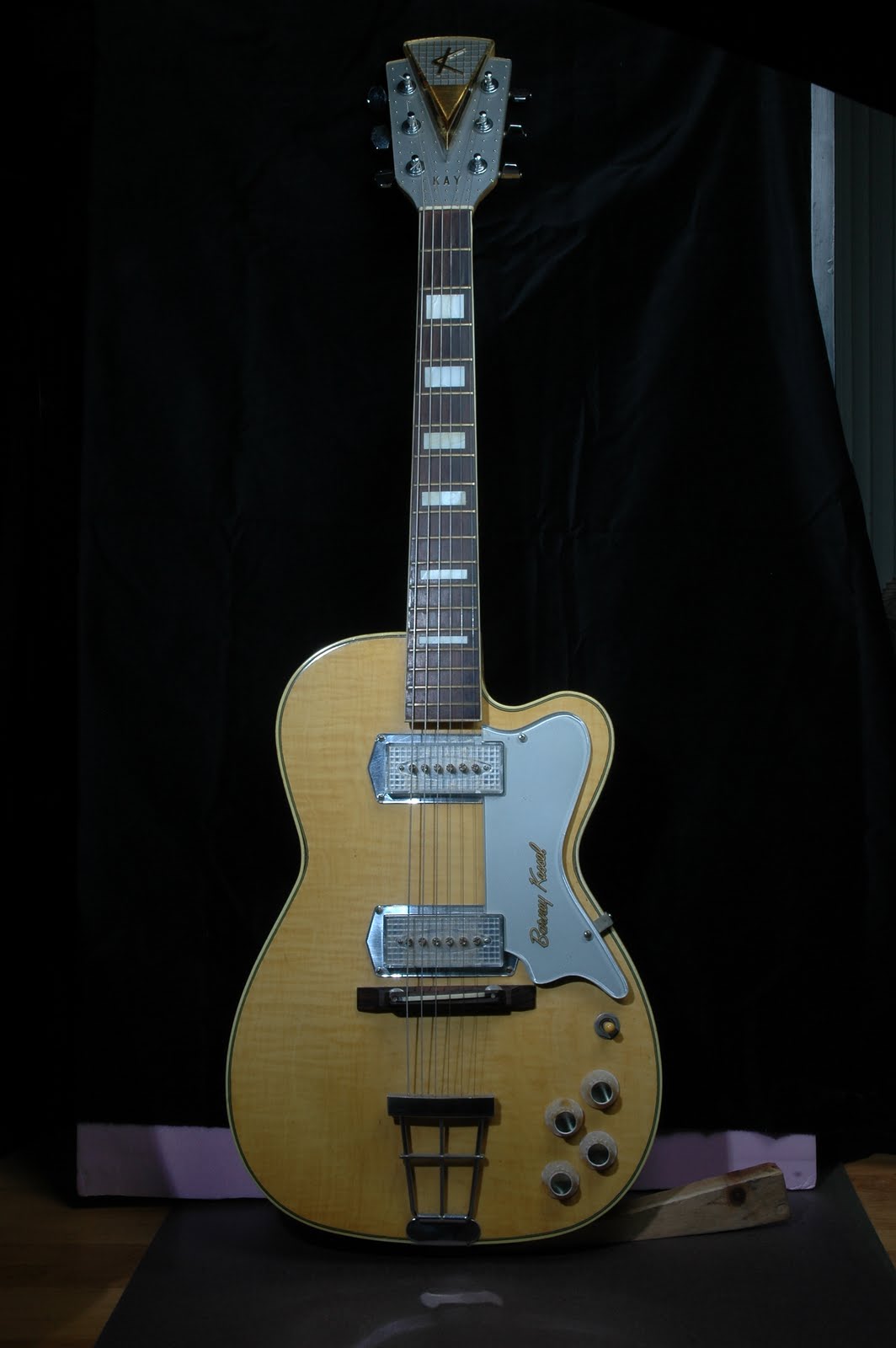 Blonde Kay Barney Kessel Pro electric guitar with two pickups and the tall Kelvinator headstock, standing against a dark curtain
